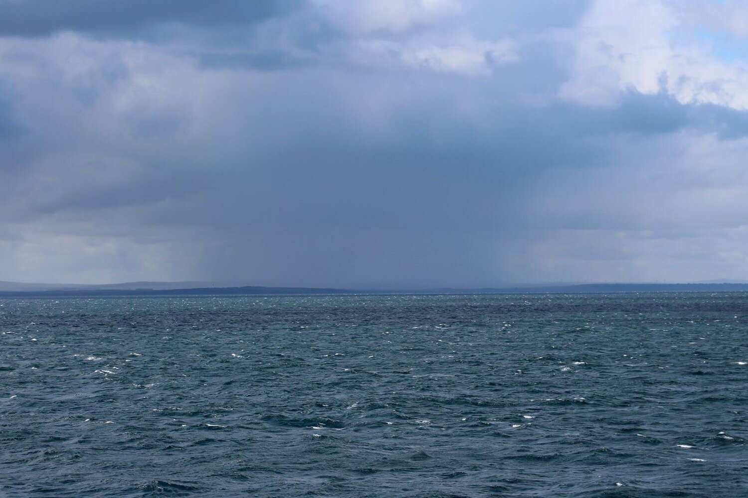 Die Nordsee ist durchquert, die Küste Schottlands kommt in Sicht. Blick auf das Meer mit dunklen Wolken und am Horizont ist Land zu sehen.
