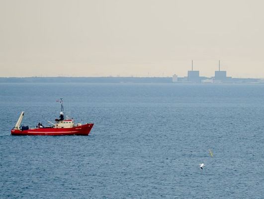 Kutter auf dem Meer. Im Hintergrund ein Atomkraftwerk an der Küste.