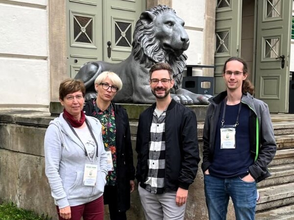 Gruppenfoto mit vier Personen, im Hintergrund ein Gebäude mit einer Löwenskulptur