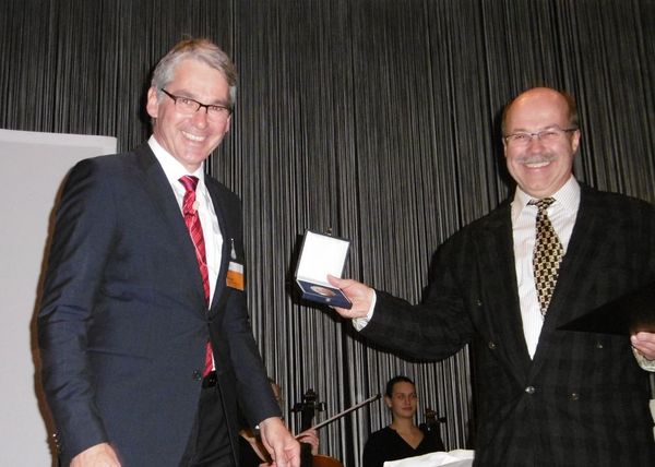 Herr Brunotte hält die Gedenkmünze in der Hand und Prof. Böttinger steht rechts von ihm daneben. Beide strahlen über das ganze Gesicht.