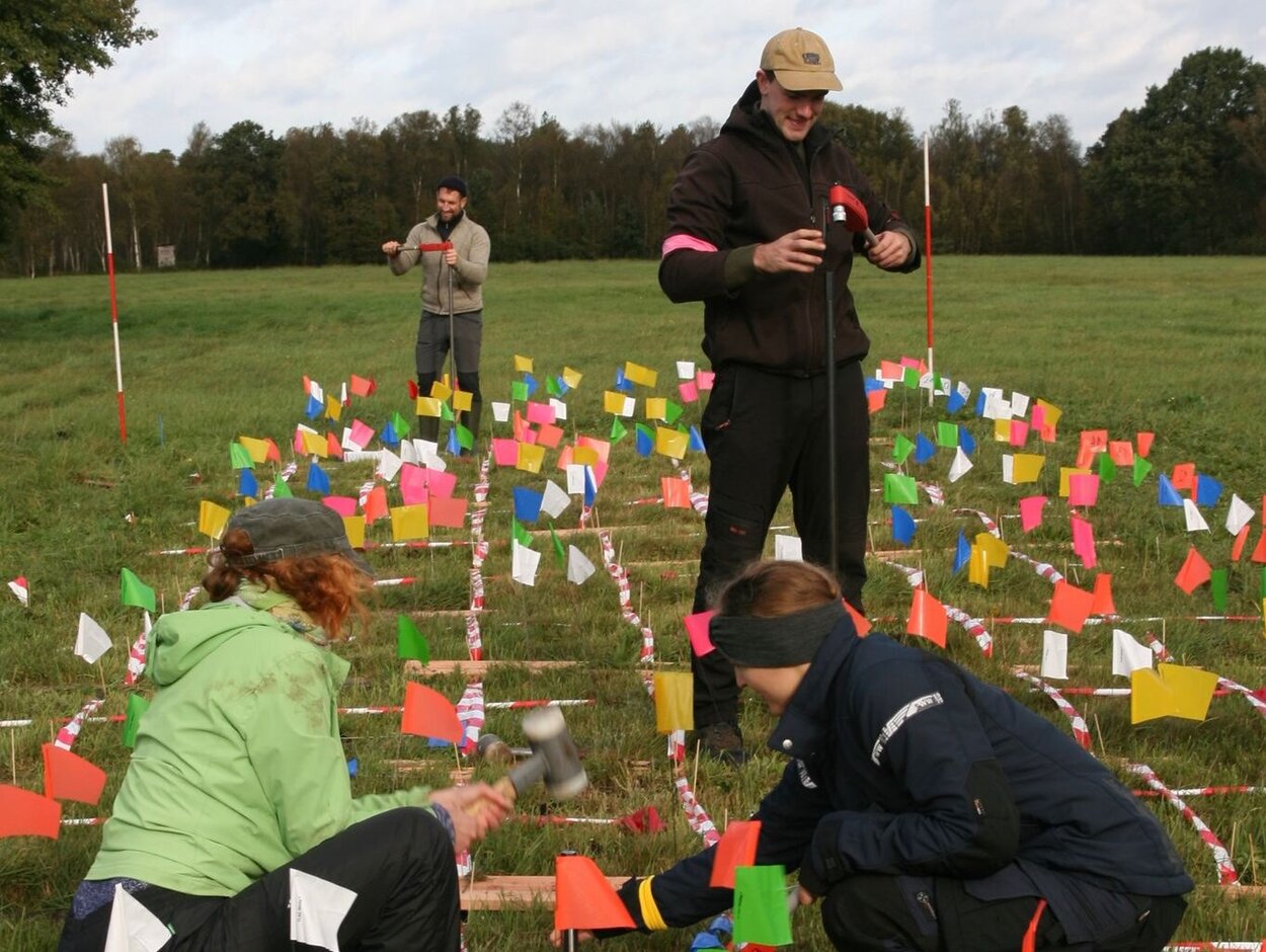Versuchsanordnung zum testen verschiedener Messinstrumente. Mehrere Personen sitzen und stehen in einem auf dem Boden marktierten Quadratfelder.