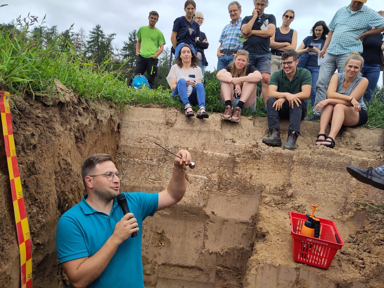 Ein Mann steht in einem ausgehobenen Bodenprofil und zeigt einem Publikum von Männern und Frauen ein Stück einer Wurzel.