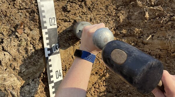 Probenentnahme Eine Person schlägt mit einem Hammer einen hohlen Zylinder in ein Bodenprofil zur Probenentnahme in einer bestimmten Tiefe.