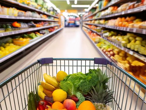 Ein mit Obst und Gemüse gefüllter Einkaufswagen in einem Supermarkt zwischen zwei Regalen.