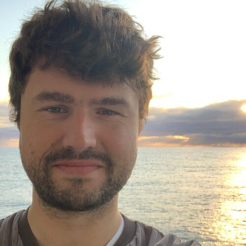 Mann blickt freundlich in die Kamera. Im Hintergrund das Meer bei Sonnenaufgang.