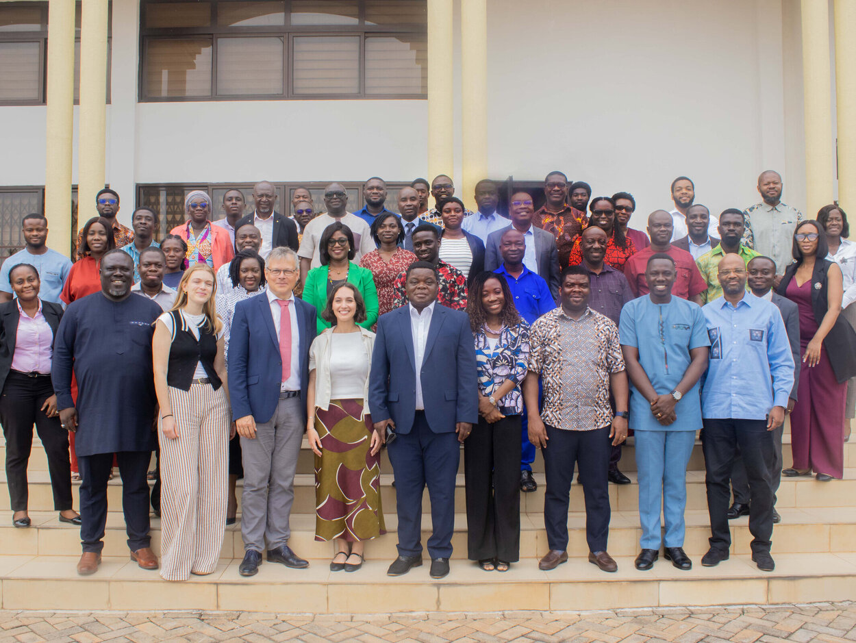 Eine große Gruppe von etwa 45 Menschen steht auf den Stufen eines Gebäudes mit hellen Säulen und Fenstern. Die Gruppe besteht aus Männern und Frauen unterschiedlicher Herkunft, viele von ihnen lächeln. Viele von ihnen tragen formelle Kleidung, einige tragen Anzüge, andere tragen traditionelle und farbenfrohe Outfits. Der Hintergrund zeigt eine weiße Wand und Glasfenster des Gebäudes.