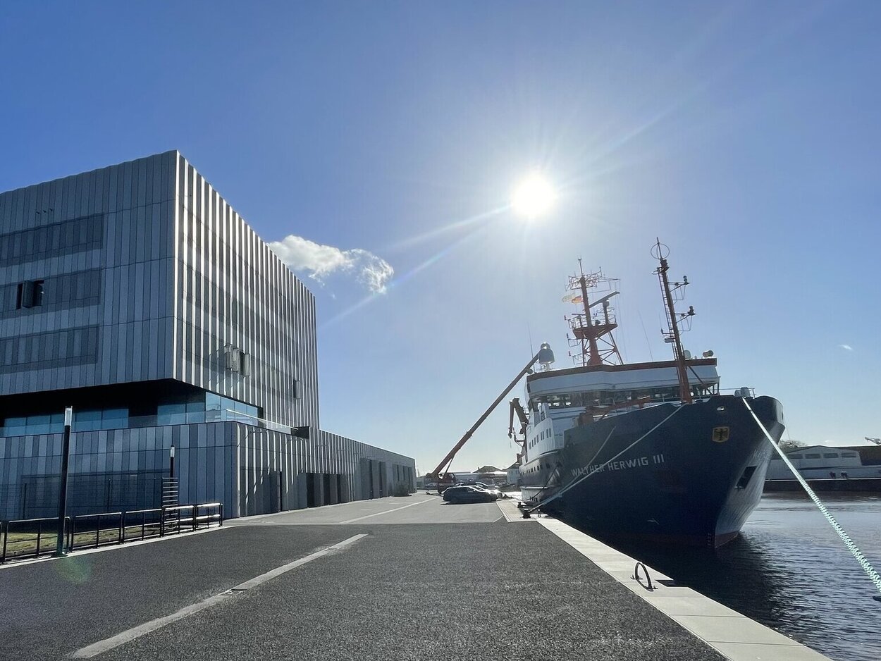 Modernes Bürogebäude neben einem Hafenbecken mit einem festgemachten Forschungsschiff unter klarem Himmel