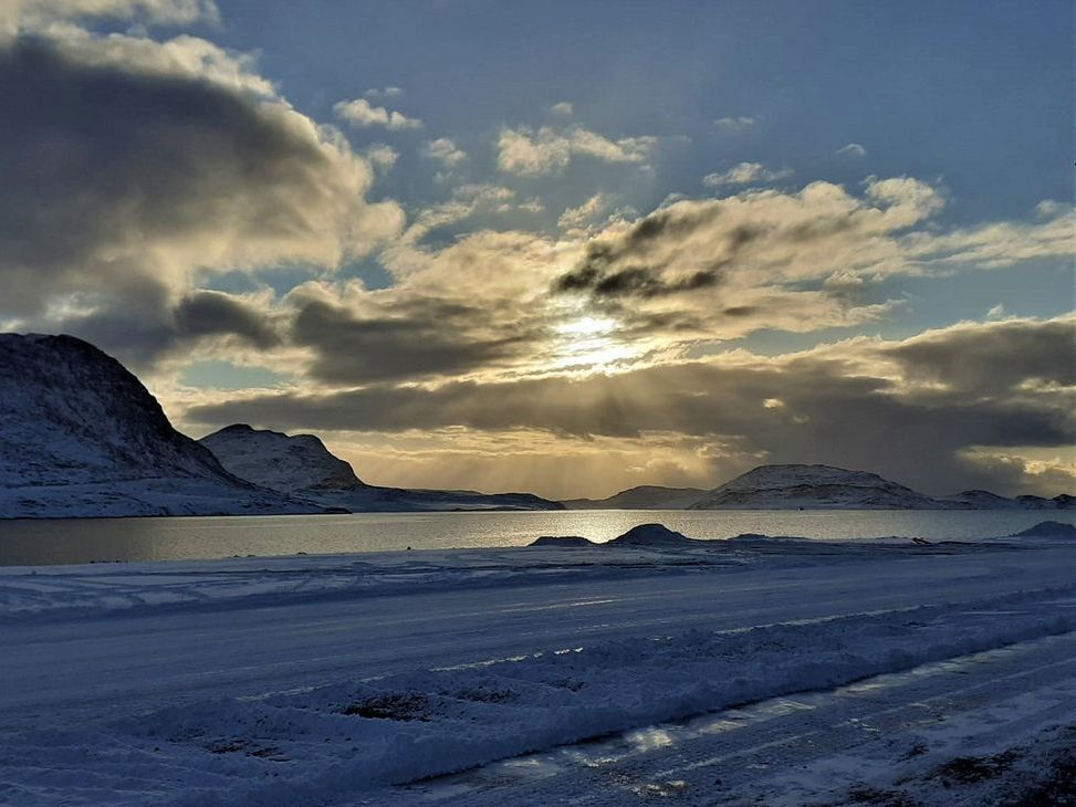 [Translate to English:] Die Landschaft auf Grönland mit wolkigem Himmel, Sonne, Bergen, Schnee und Wasser.