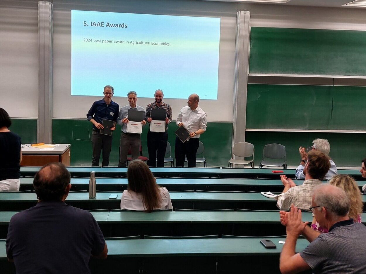 Preisträger auf der EAAE. Vier Personen stehen vor einer Leinwand in einem Saal und Zuschauer applaudieren.