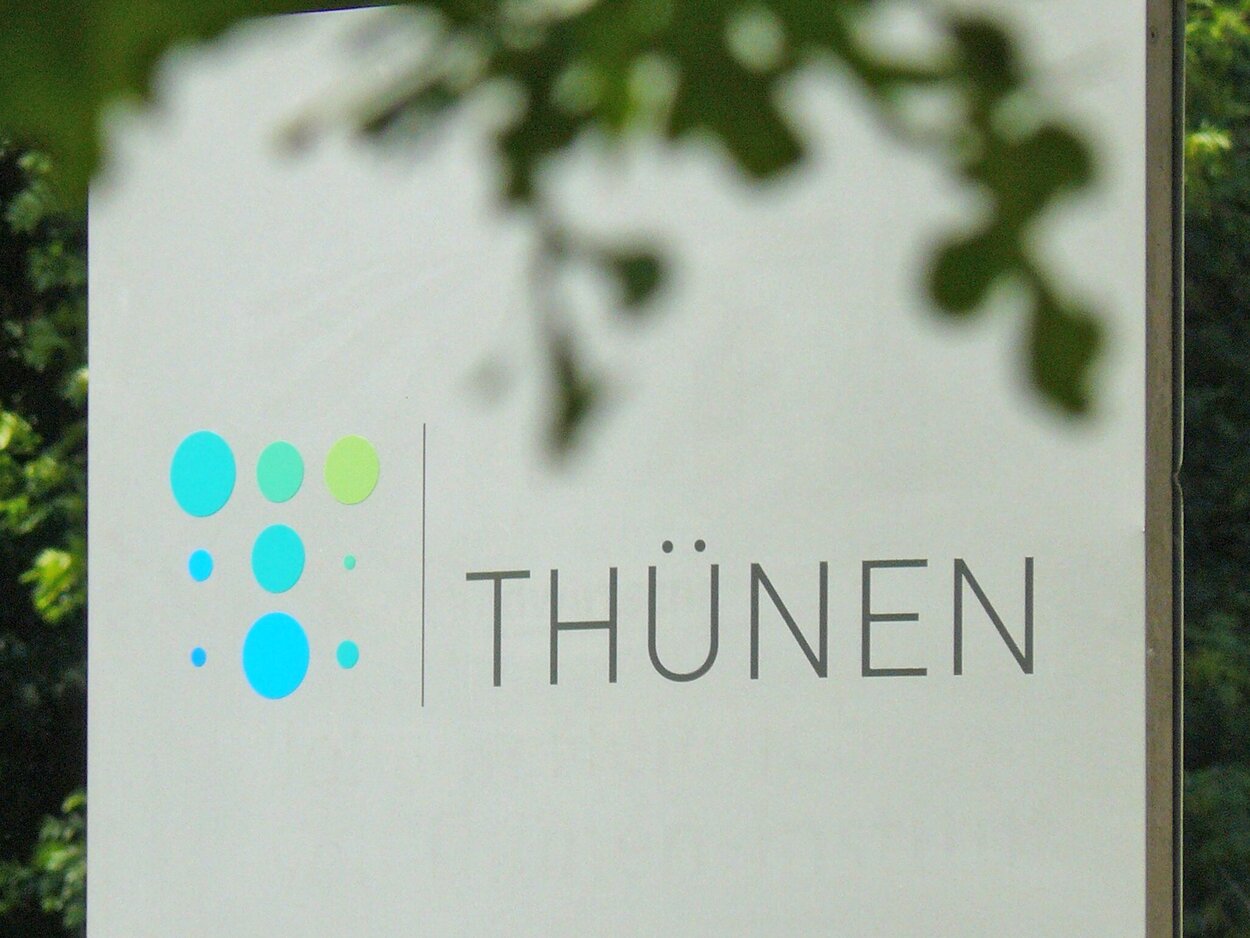 Eine Metallstele mit dem Logo des Thünen-Instituts mit Bäumen im Vorder- und Hintergrund.
