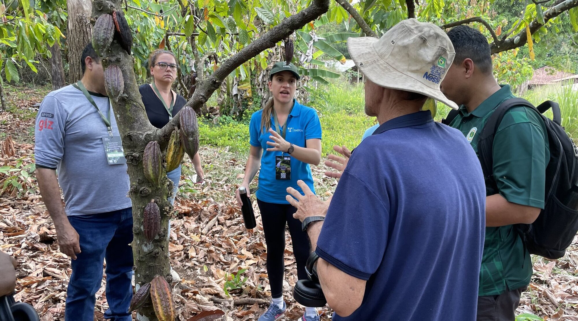 Caroline Salomão diskutiert die Auswirkungen der neuen EU Verordnung mit lokalen Kakaoproduzenten und Vertreter*innen der Regierung und Nichtregierungsorganisationen. Mehrere Personen stehen um einen Baum.
