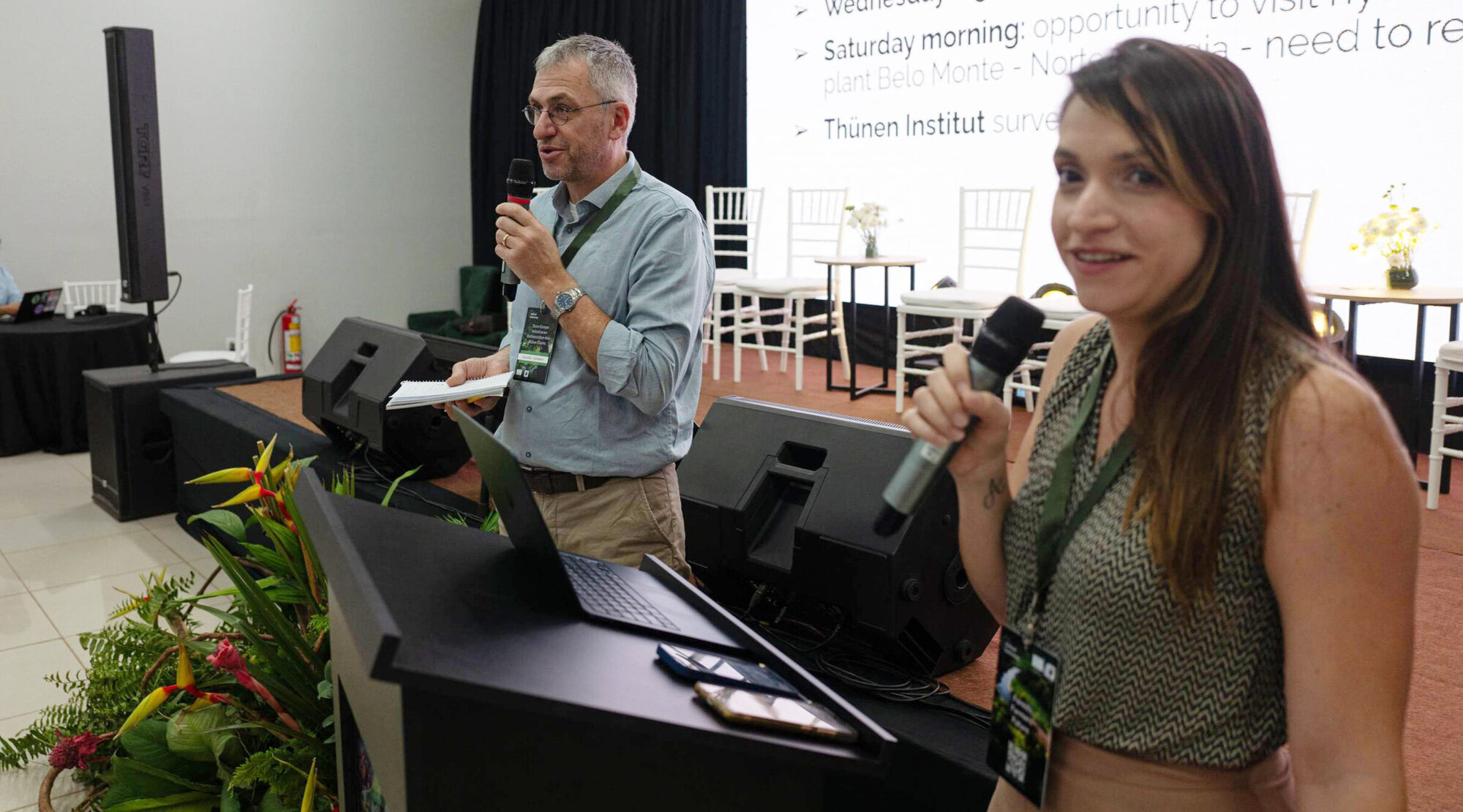 Richard Fischer und Caroline Salomão eröffnen eine interaktive Befragung der Konferenzteilnehmer zu den Auswirkungen der EUDR. Zwei Personen stehen an einem Rednerpult mit Mikofonen in der Hand und schauen in die Kamera.