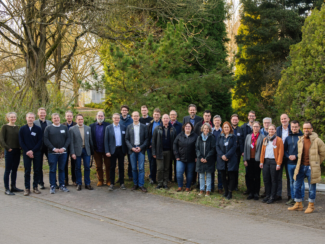Eine Gruppe von 28 Menschen auf dem Außengelände eines Forschungsinstitutes. Hinter den Menschen wachsen Bäume, dahinter ist ein großes Gewächshaus zu sehen.