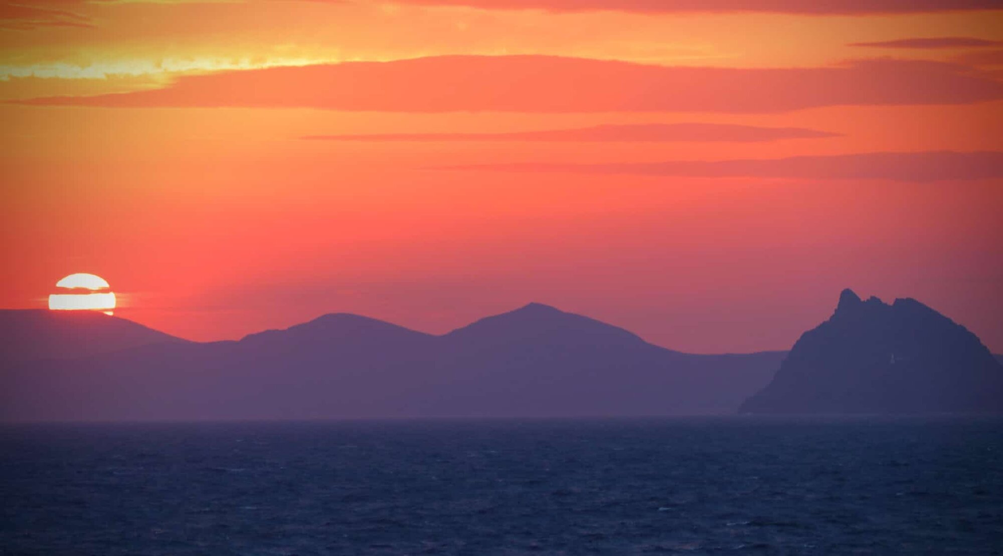 Sunrise off the Irish coast. A dark sea with dark mountains and a red sky with veiling clouds.