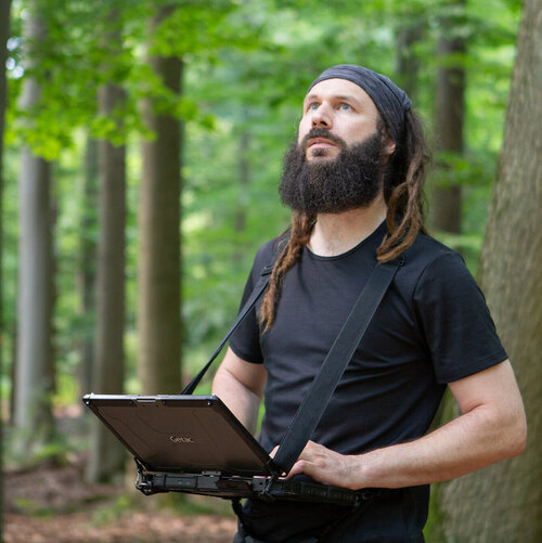 Mann mit Bart und Stirnband im Wald hält ein offenes, Notebook-ähnliches Gerät und schaut nach oben.