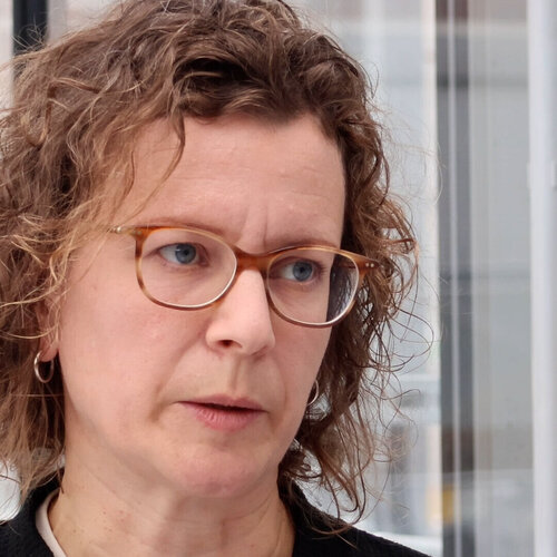 Porträt einer Person mit braunen, gelockten Haaren und Brille, die spricht.