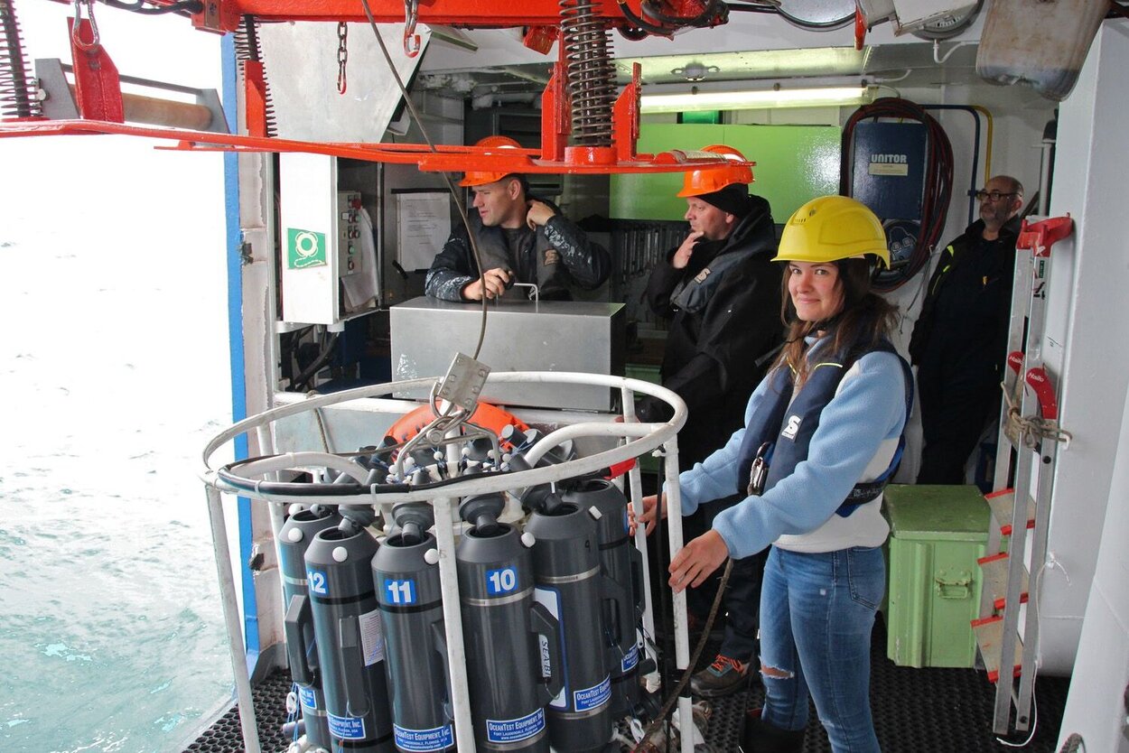 Die CTD-Rosette wird zum Aussetzen vorbereitet. Eine Forscherin mit gelbem Helm und Rettungsweste lässt ein tonnenformiges, ringsum mit geöffneten Röhren bestücktes Messinstrument zu Wasser. Zwei Personen im Hintergrund bedienen die Winde.