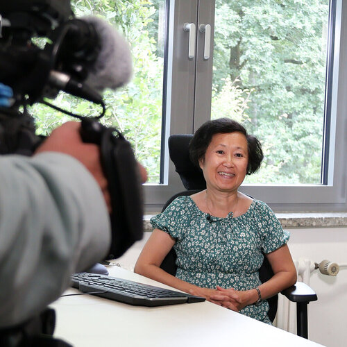 Frau sitzt an einem Schreibtisch mit Tastatur, trägt ein grünes, geblümtes Kleid und sitzt vor einem Fenster mit Blick auf Bäume. Im Vordergrund eine professionelle Kamera, die auf sie gerichtet ist.