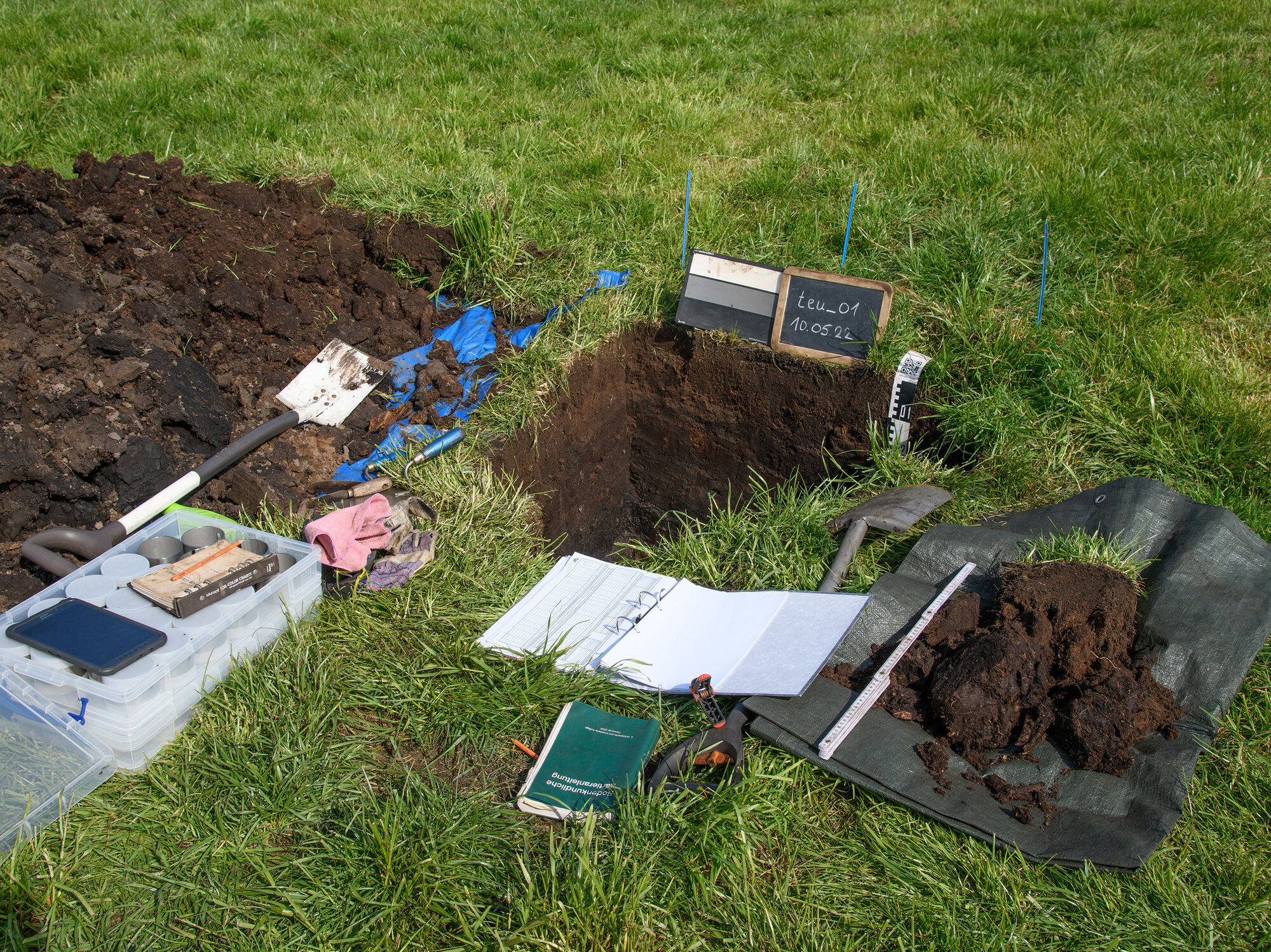 Anhand von Profilgruben beschreiben die Kartierteams die Moorböden. Auf einer Wiese ist eine Grube ausgehoben und es liegen Papier und Schaufel bereit.