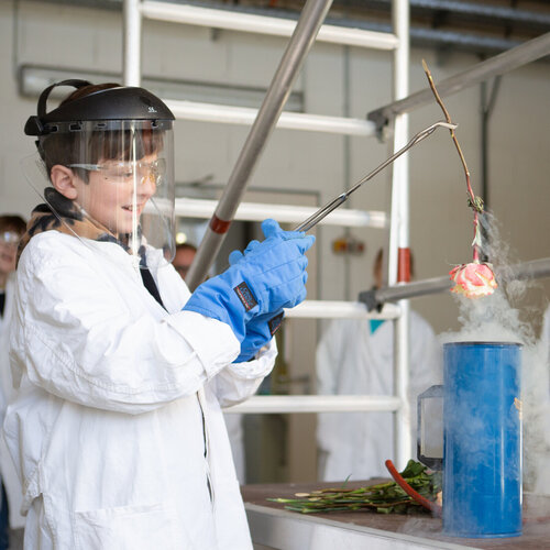 Ein Junge im Laborkittel und Gesichtschutz hält mit einer Zange eine Rose über einem langen, blauen Glasgefäß. Aus dem Gefäß steigt weißer Dampf auf.